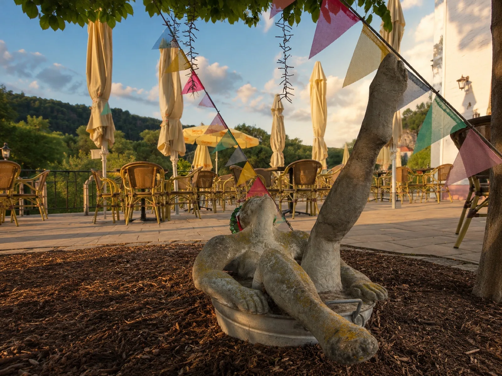 Steinskulptur im Garten der Mühle, dahinter die Terrasse im Abendlicht mit bunten Wimpeln